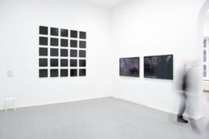 Eine minimalistische Kunstgalerie mit weißen Wänden zeigt 25 kleine, gleichmäßig verteilte schwarze quadratische Rahmen, die in einem Raster an der linken Wand angeordnet sind. Zwei größere rechteckige Kunstwerke hängen an der angrenzenden Wand. Eine verschwommene Gestalt geht über den grauen Boden und natürliches Licht fällt durch ein Fenster ein. (KI generierte Beschreibung)