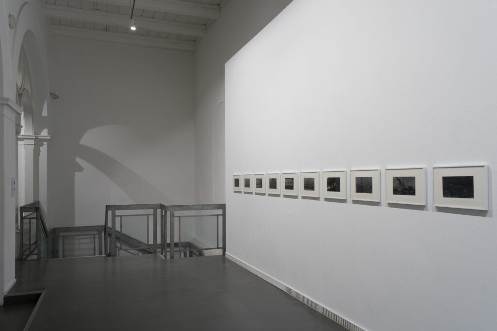 Eine minimalistische Kunstgalerie zeigt eine Reihe gerahmter Fotografien, die gleichmäßig auf einer weißen Wand verteilt sind. Der Boden der Galerie ist dunkel und glatt und bildet einen Kontrast zu den weißen Wänden. Ein Geländer mit Glasplatten grenzt an eine Treppe oder eine untere Ebene, und eine gewölbte Decke sorgt für architektonisches Interesse. (KI generierte Beschreibung)