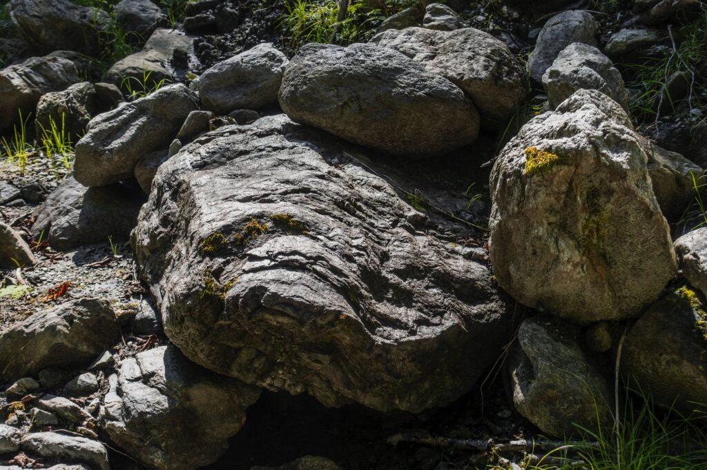 Eine Ansammlung großer, verwitterter Felsen, die teilweise mit Moos bedeckt und von verstreutem Gras und kleinen Pflanzen umgeben sind. Die Felsen haben eine ungleichmäßige Textur und Größe. Das Sonnenlicht wirft Schatten und hebt die natürliche Landschaft und die Rauheit der Umgebung hervor. (KI generierte Beschreibung)