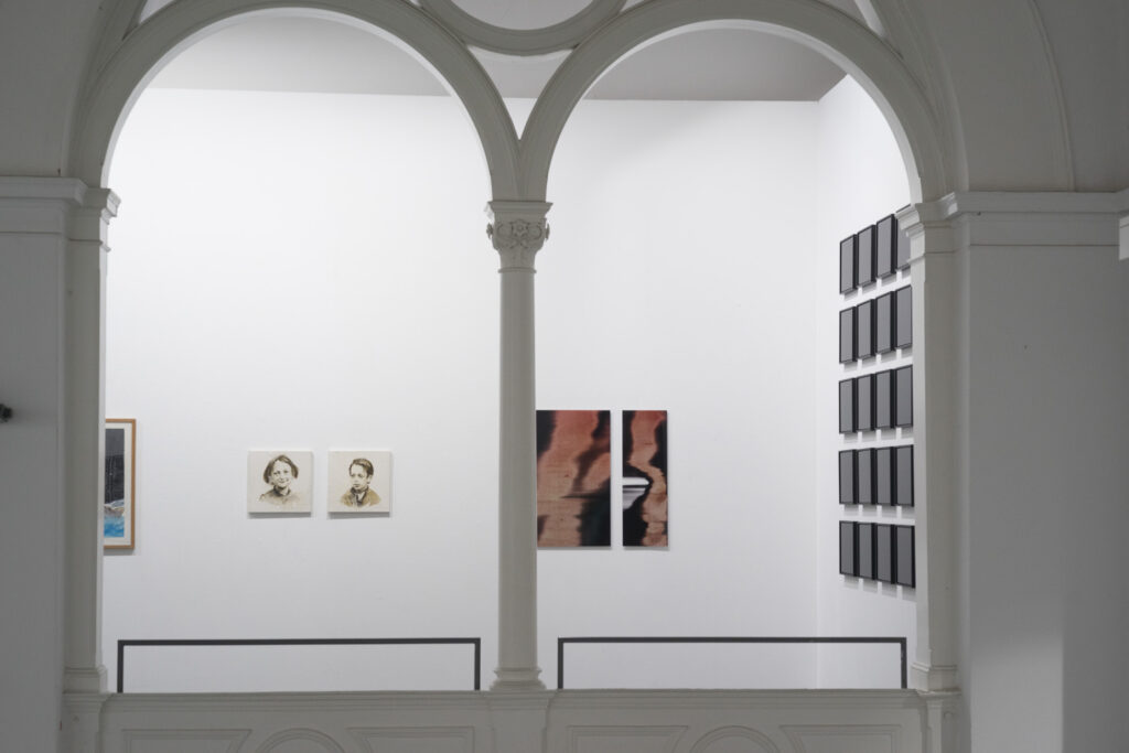 Blick auf eine Kunstgalerie mit zwei großen weißen Torbögen, die mehrere Kunstwerke an der Wand einrahmen. Die Ausstellung umfasst zwei Porträts, ein abstraktes Werk mit verzerrten Linien und ein Raster aus kleinen schwarzen rechteckigen Rahmen, die in Spalten angeordnet sind, alles vor einem strahlend weißen Hintergrund. (KI generierte Beschreibung)