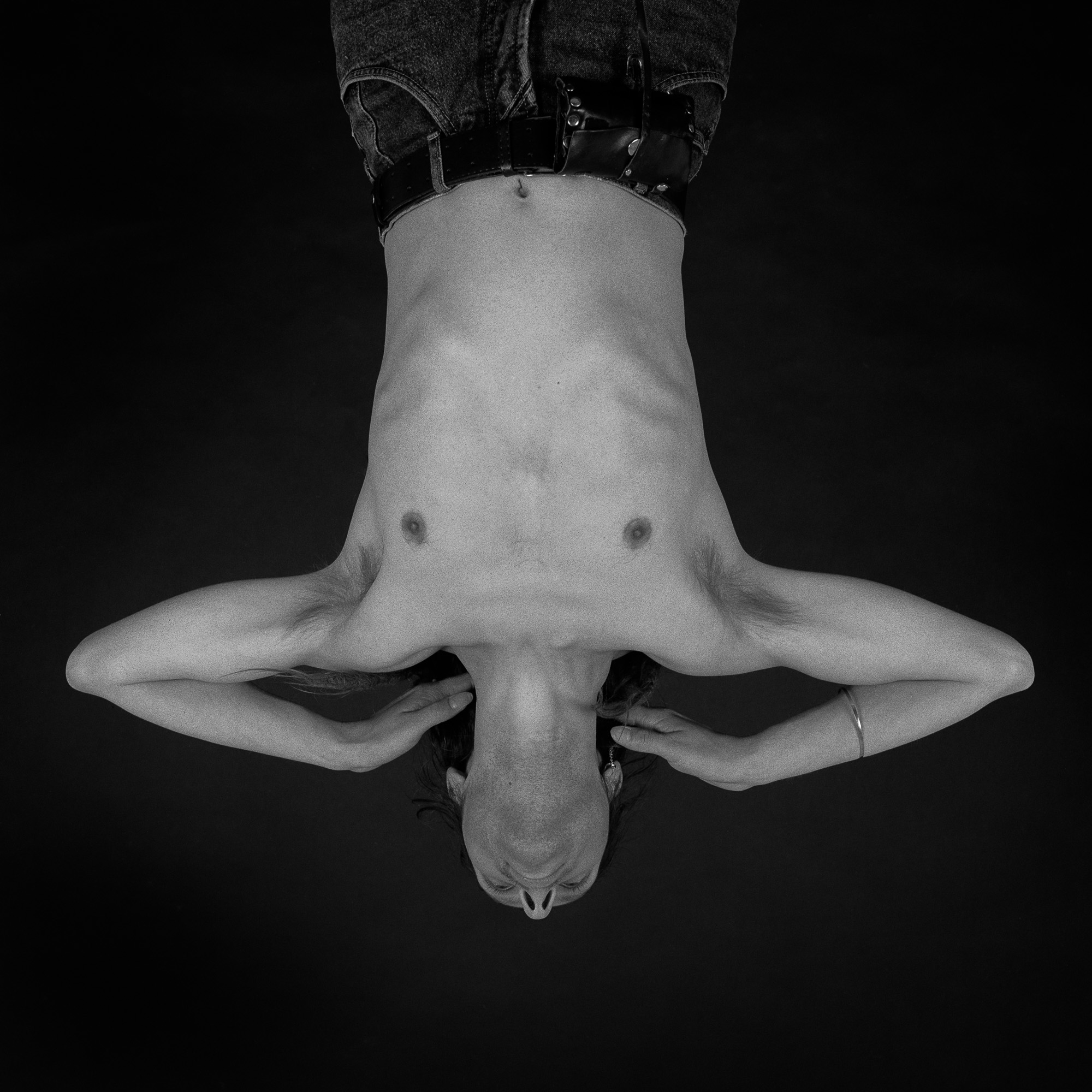 Ein Schwarz-Weiß-Foto zeigt eine Person ohne Hemd, die mit angewinkelten Armen und den Händen am Kopf auf dem Rücken liegt. Das Bild ist von oben aufgenommen; Jeans und Gürtel sind sichtbar. Der dunkle Hintergrund betont die Konturen und die Haltung des Körpers. (KI generierte Beschreibung)