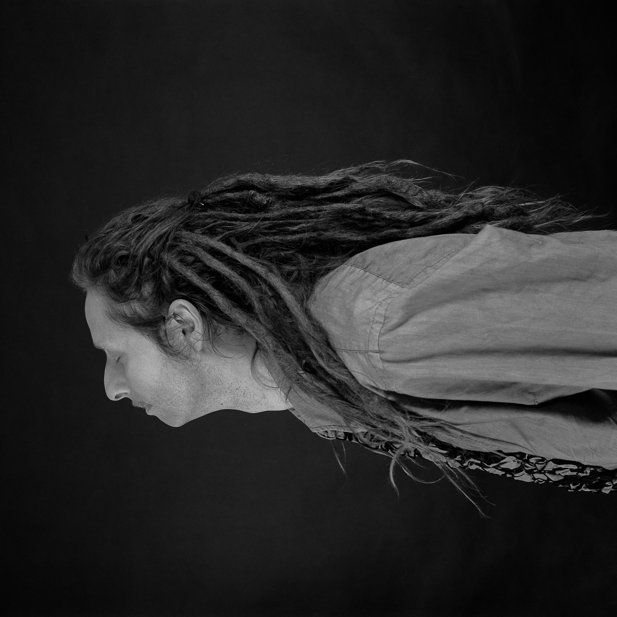Eine Person mit langen, dichten Dreadlocks ist im Profil vor dunklem Hintergrund fotografiert. Sie blickt nach links und trägt ein helles Hemd und ein gemustertes Kleidungsstück oder einen Schal. Das Bild ist schwarz-weiß und betont Textur und Kontrast. (KI generierte Beschreibung)