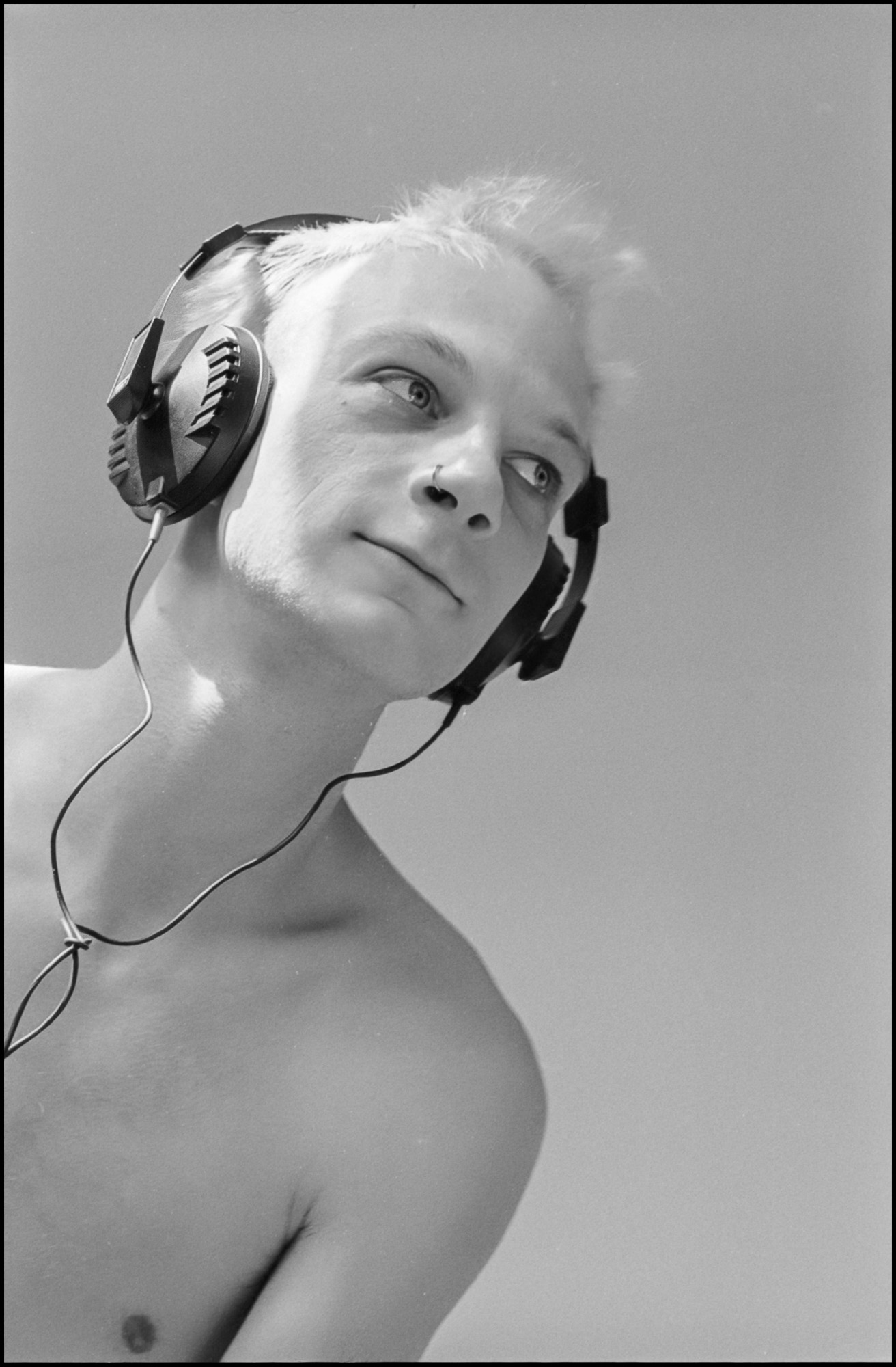 Eine junge, hemdlose Person mit kurzem, hellem Haar trägt große Over-Ear-Kopfhörer und blickt mit einem leichten Lächeln nach oben. Der Hintergrund ist schlicht und hell beleuchtet, was die entspannte und nachdenkliche Stimmung unterstreicht. Das Bild ist in schwarz-weiß gehalten. (KI generierte Beschreibung)